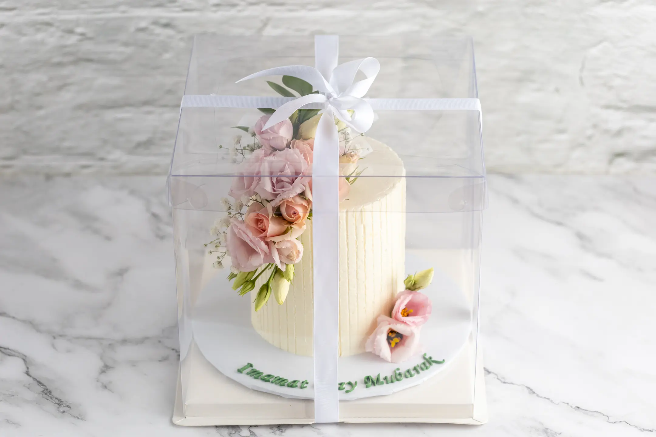ALT: Single-tier round cake with vertical textured finish and pastel pink sugar flowers in clear box with white ribbon CAPTION: A delicate single-tier cake with soft florals and textured finish, elegantly presented in a clear gift box. Design yours. DESCRIPTION: This single-tier round cake features a subtle vertical textured finish in a creamy hue, adorned with a cluster of pastel pink sugar flowers and green leaves. The design is refined and gentle, perfect for intimate celebrations or thoughtful gifts. Presented in a clear box tied with a white ribbon, it offers a polished and sophisticated presentation. Customise the floral arrangement or finish to suit your occasion. Crafted with care by Sucré in Toronto.