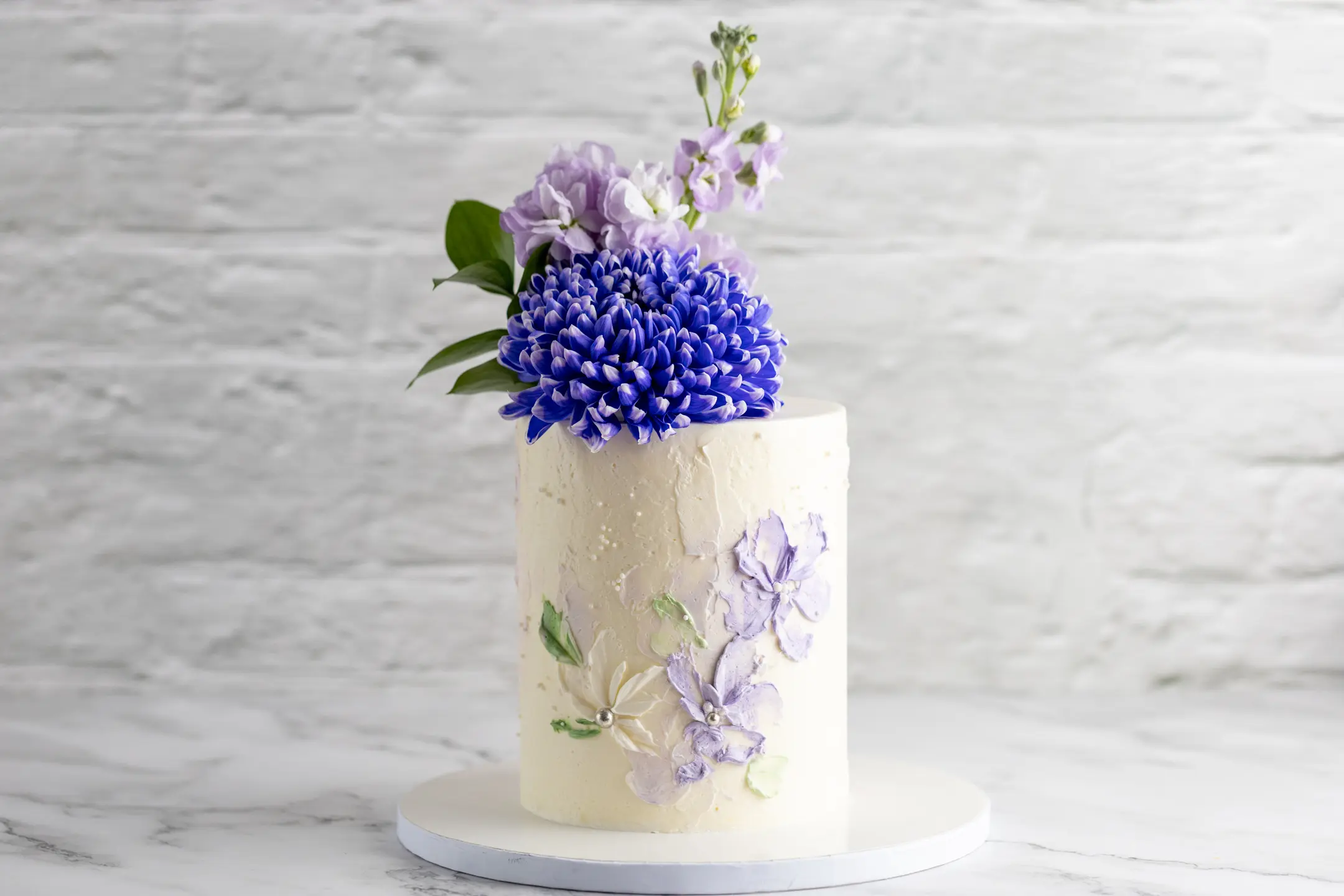 ALT: Single-tier cylindrical cake with textured white finish, purple and white floral painting, and fresh purple flowers topper CAPTION: A refined single-tier cake with delicate floral painting and fresh blooms for a natural, elegant touch. Design yours. DESCRIPTION: This single-tier cylindrical cake features a textured white finish, enhanced by hand-painted purple and white flowers with subtle green leaves. The design is crowned with a fresh floral arrangement in shades of purple, adding a vibrant yet soft contrast. The combination of painted and fresh florals creates a harmonious balance of artistry and nature. Perfect for intimate celebrations or as a sophisticated statement piece, this cake exemplifies Sucré’s commitment to craftsmanship and timeless elegance. Custom options are available to suit your event’s palette and style.