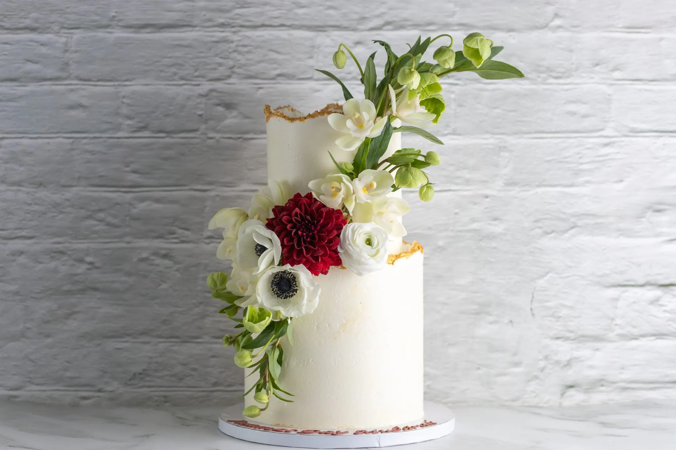 ALT: Two-tier round cake with smooth white finish, gold leaf edges, and fresh white and red floral arrangement CAPTION: A refined two-tier cake with fresh florals and delicate gold leaf accents. Design yours with Sucré. DESCRIPTION: This elegant two-tier round cake features a smooth white finish accented by subtle gold leaf along the edges. A fresh floral arrangement cascades naturally down the side, combining white blooms with a striking deep red flower for contrast. The design balances simplicity with thoughtful detail, making it suitable for weddings or special celebrations. Crafted with care by Sucré in Toronto, this cake invites customization in flowers, colours, and tier proportions to perfectly complement your event.