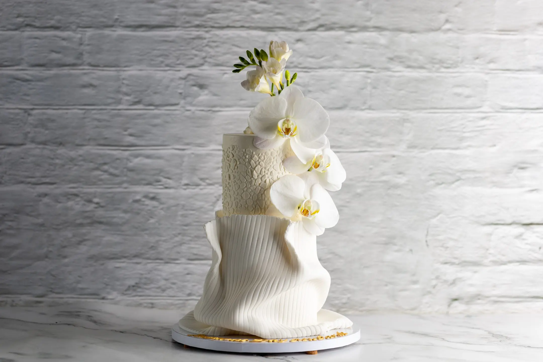ALT: Two-tier round white cake with textured finish and draped fondant, adorned with white orchids and freesia CAPTION: A sculptural white cake featuring delicate orchids and elegant draping. Design yours with Sucré. DESCRIPTION: This two-tier round cake showcases a refined blend of textures, with a softly embossed upper tier and a flowing draped fondant on the lower tier. The design is accented by fresh white orchids and freesia, adding natural elegance and a touch of organic beauty. The neutral palette and sculptural form create a modern yet timeless aesthetic, perfect for weddings or sophisticated celebrations. Crafted with care by Sucré in Toronto, this cake invites personal touches to suit your special occasion.