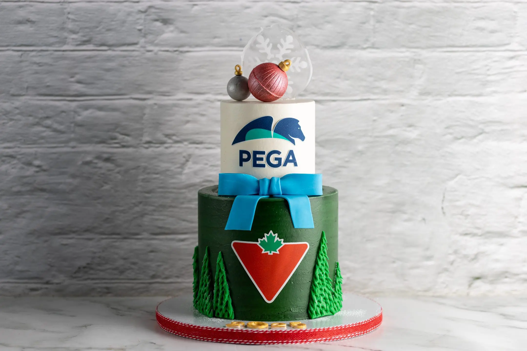 ALT: Two-tier round cake with green textured lower tier, white upper tier with logo, blue bow, and festive topper CAPTION: A refined two-tier design blending corporate logos with festive accents and a crisp blue bow. Design yours. DESCRIPTION: This two-tier round cake features a deep green textured lower tier adorned with a red and white logo and piped evergreen trees. The smooth white upper tier showcases a bold blue logo, accented by a striking blue fondant bow at the base. Topped with delicate festive ornaments and a translucent snowflake, the cake balances corporate branding with seasonal charm. Perfect for holiday corporate events or celebrations, this design reflects Sucré’s attention to detail and elegant craftsmanship. Custom options available to suit your occasion.